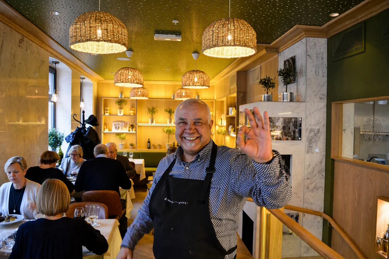 Verten i sort forkle vinker mot kamera midt i salen